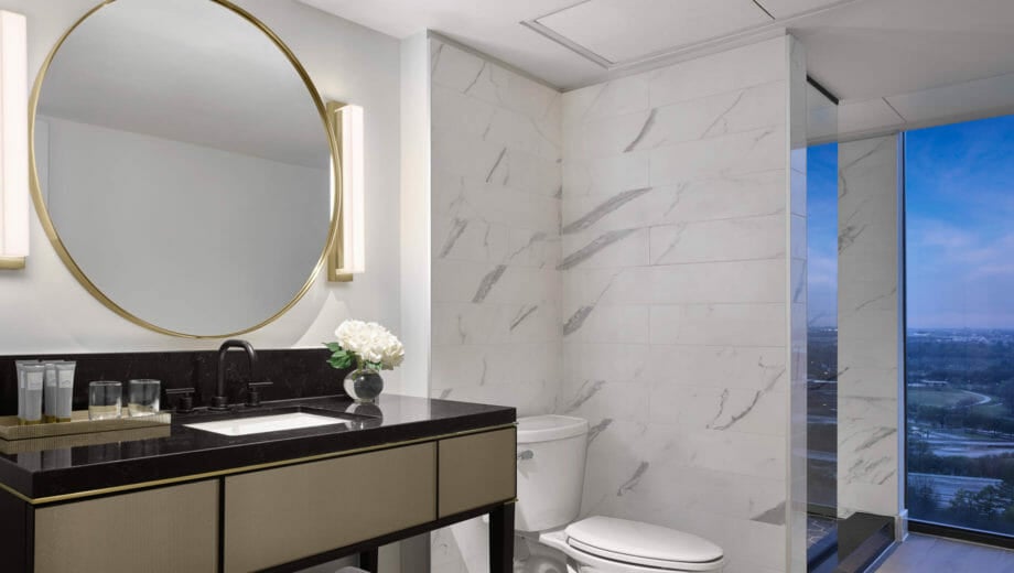 Bathroom vanity with gold framed circular mirror, marble wall tile, and entry to bathroom with floor to ceiling windows.