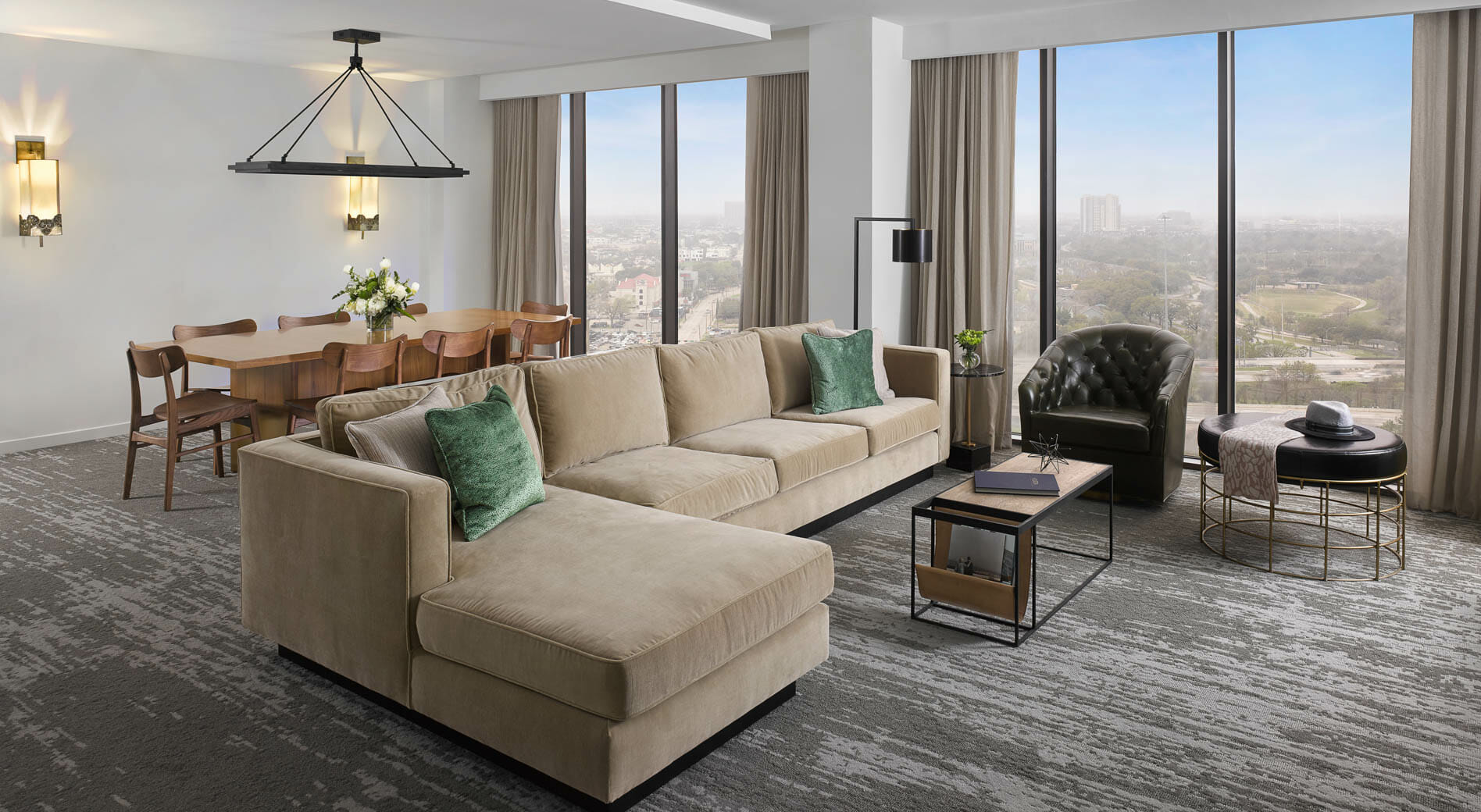 Spacious Presidential Suite living room with floor to ceiling windows, tan sectional in seating area and dining table behind it.