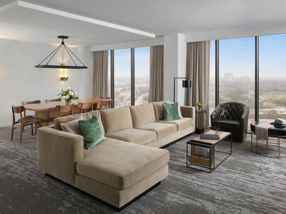 Spacious Presidential Suite living room with floor to ceiling windows, tan sectional in seating area and dining table behind it.