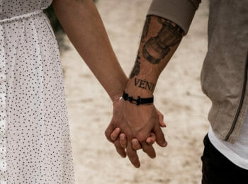 A man and woman holding hands. The man has tattoos on his arm and is wearing a gray jacket and white shirt, the woman is wearing a white dress with black polkadots.