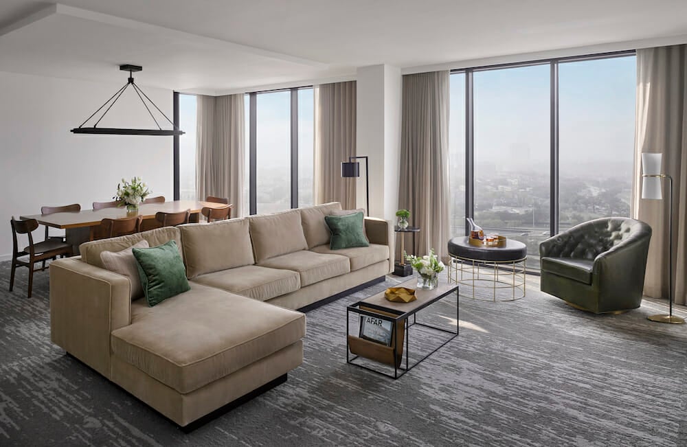 Spacious Presidential Suite living room with floor to ceiling windows, tan sectional in seating area and dining table behind it.